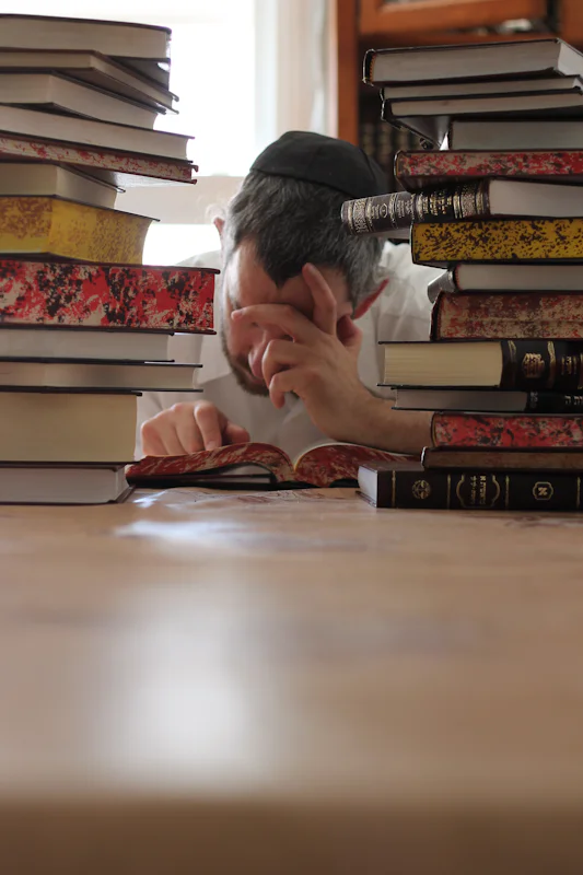 A man studying from an open book between stacks of books